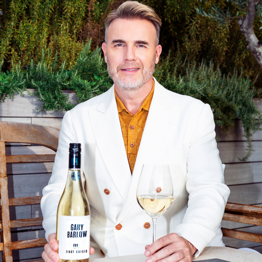 Gary Barlow holding a glass of Pinot Grigio with the wine bottle on the table, showcasing his wine in a relaxed setting.