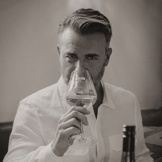 Black and white image of Gary Barlow enjoying a glass of Sauvignon Blanc, wearing a white shirt at a dining table with a wine bottle in the foreground.