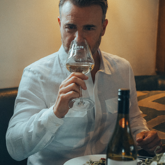 Gary Barlow enjoying a glass of Gary Barlow Sauvignon Blanc at a dining table, elegantly dressed in a white shirt, with a bottle of Sauvignon Blanc in the foreground.