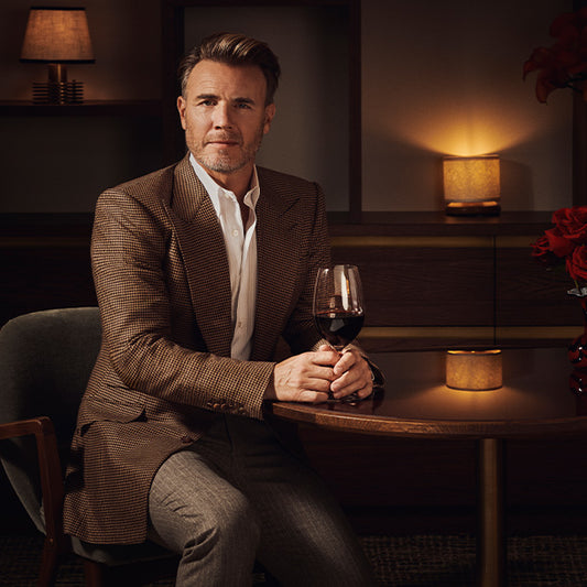 Gary Barlow seated at a luxurious table, elegantly holding a glass of red wine from his wine collection, set in a warmly lit, stylish ambiance.